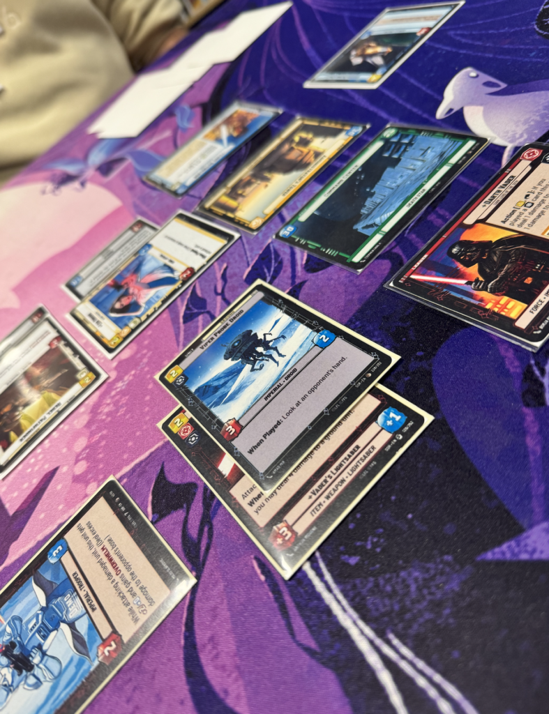 Screenshot 2026-04-26 at 2.32.53 PM The image shows several trading card game playing cards laid out on a purple playmat. The playmat features abstract and possibly fantasy-themed designs, with swirling patterns and a bird-like figure on the right. The cards are variously positioned, some overlapping, showcasing different illustrations and colors. Cards visible include imagery like landscapes and characters, with varied borders indicating their types or rarities. There is a presence of a hand or arm on the left side, suggesting a player is involved in the game.
